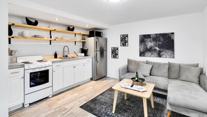 A modern kitchen with white cabinets, a stainless steel refrigerator, and a built-in oven. There is a grey sofa and a small wooden table with a bottle and glasses. - Spacious 1BR Near Downtown & Water w/ Full Kitchen (Lake Worth)