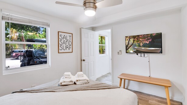 A bedroom with a bed, a ceiling fan, a TV, a bench, and a door leading to another room. - Spacious 1BR Near Downtown & Water w/ Full Kitchen (Lake Worth)