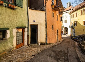 Una stretta strada acciottolata fiancheggiata da edifici storici, con un'architettura variegata e un cartello che indica uno spazio pubblico.