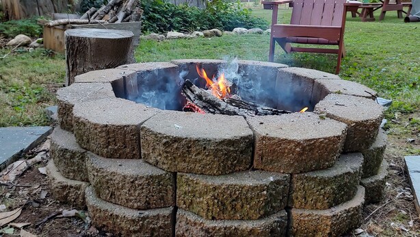 Un pozo de fuego circular de piedra con un fuego ardiendo en su interior, rodeado por un césped donde se ve una casa de madera y una silla roja al fondo.