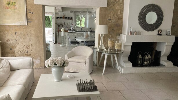 Un salón con un sofá blanco, una mesa de centro, un juego de ajedrez, un jarrón con flores, una chimenea y una cocina al fondo.