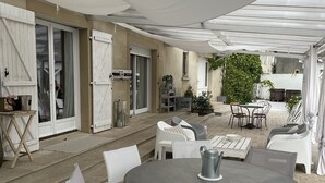 Un patio al aire libre con una mesa redonda y sillas blancas, una pérgola sobre la cabeza y una puerta que da a un vestíbulo cubierto.