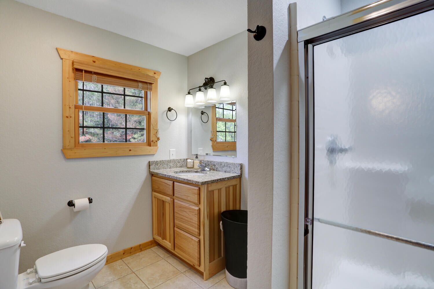 A bathroom with a toilet, wooden vanity, sink, and a window with blinds.