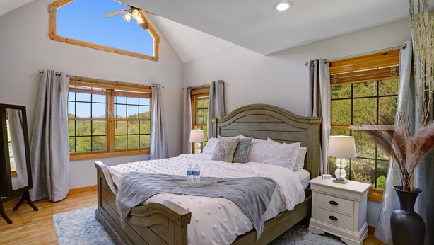 A bedroom with a large bed, a nightstand, a mirror, and a window with a view of greenery. - NEW! Valley View Mountain Getaway (Hiawassee)