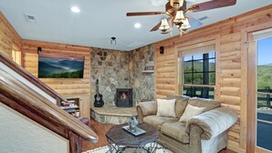 A cozy log cabin interior with a stone fireplace, a comfortable sofa, a guitar, and a television mounted on the wall. - NEW! Valley View Mountain Getaway (Hiawassee)