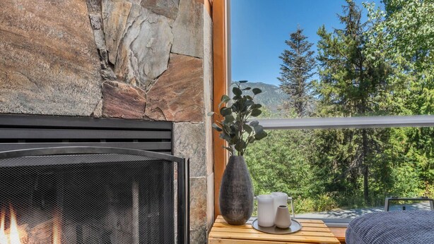 A stone fireplace with a metal screen, a wooden side table with a vase and candles, and a view of trees and mountains through a large window. - 3BR | Whistler Creek Ridge | Ski-in/out | Private Hot Tub | Wood Fire | Private Garage (Whistler)