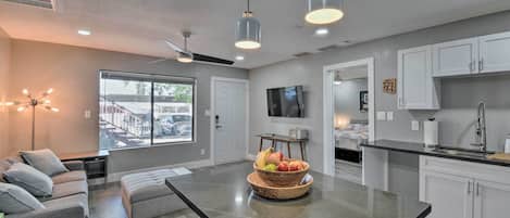 A modern kitchen with white cabinets, a large island with a fruit bowl, and a dining area with a sofa and a coffee table.