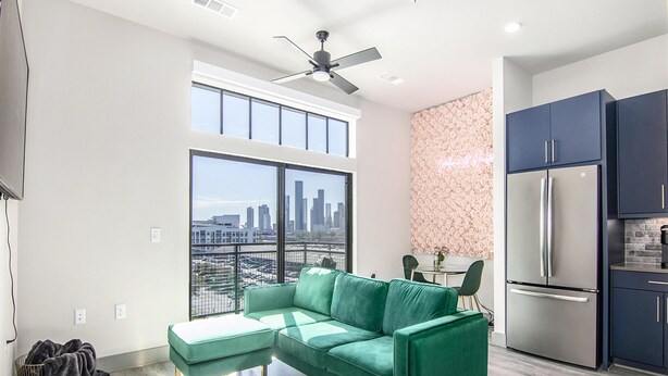 A modern apartment with a green sofa, a dining area, a kitchen with a stainless steel refrigerator, and a city view through large windows.