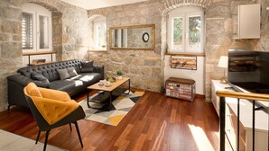 A living room with a stone wall, a black sofa, a mustard-colored armchair, a wooden coffee table, and a television. - Sun Spalato Giardino (Split)