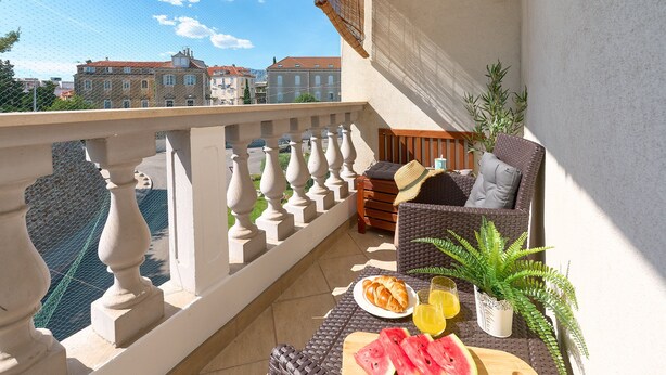 A balcony with wicker furniture, a dining table with food and drinks, and a view of buildings in the background. - Sun Spalato Giardino (Split)
