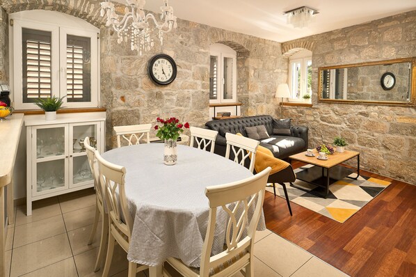 A dining area with a round table, chairs, and a chandelier. There is a stone wall with a clock and a mirror.