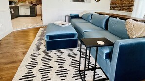 Een moderne woonkamer met een blauwe modulaire bank, een klein bijzettafeltje en een kamerkleed met een patroon. Er is een keuken zichtbaar op de achtergrond, met een bakstenen muur en een hangspiegel.