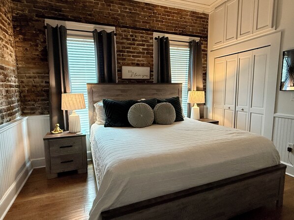 Un dormitorio con pared de ladrillo, cabecera de madera, cama con ropa de cama blanca, dos mesitas de noche con lámparas y una ventana con persianas.