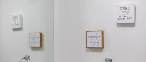 Un baño con un perchero, un cartel que dice '¿Necesitas papel higiénico? Mándame un mensaje' y un cartel que dice 'ESTE BAÑO ES PARA cantar'.