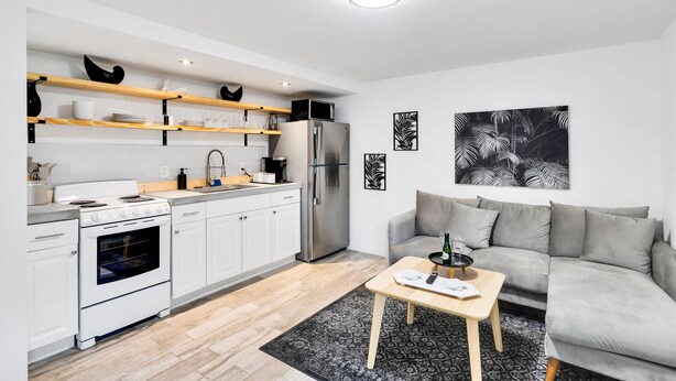 Een moderne keuken met witte kasten, een roestvrijstalen koelkast en een inbouwoven. Er staat een grijze bank en een kleine houten tafel met een fles en glazen.