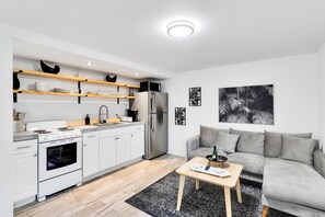 A modern kitchen with white cabinets, a stainless steel refrigerator, and a built-in oven. There is a grey sofa and a small wooden table with a bottle and glasses. - Cute 2BR Condo Steps to Waterfront Park w/ SmartTV (Lake Worth)