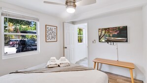 Een slaapkamer met een bed, een plafondventilator, een televisie, een bankje en een deur naar een andere kamer.