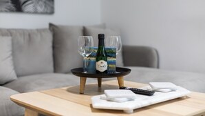 Een tafel met een fles wijn, twee glazen, een afstandsbediening en een onderzetter, in een woonkamer met een grijze bank en een schilderij op de achtergrond.