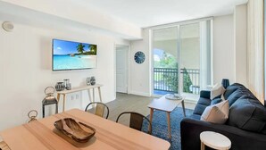 A modern living room with a large TV displaying a beach scene, a dining area with a wooden table, and a sofa with cushions.