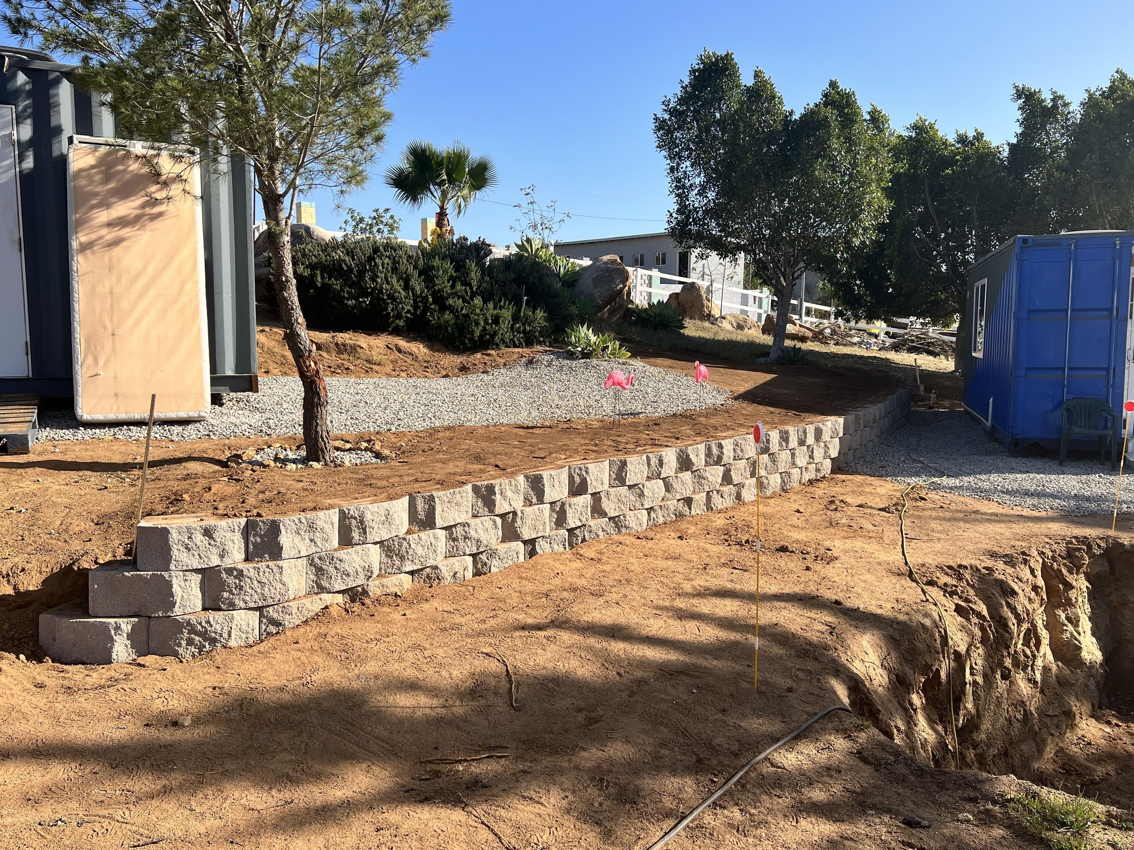 A construction site with a stone retaining wall, a small tree, and a blue container.