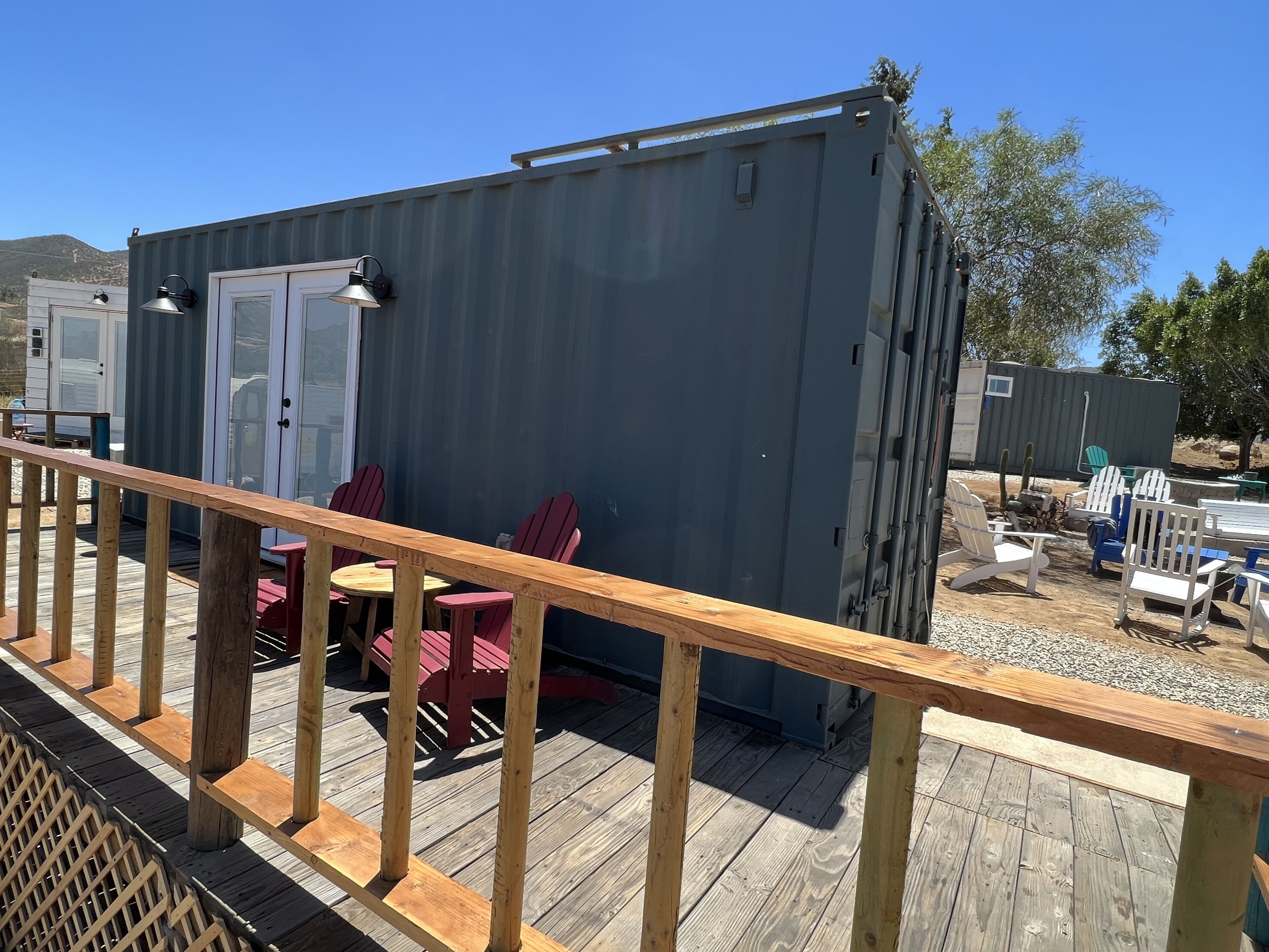Une structure compacte en métal gris, dotée d’une porte et de fenêtres, jouxtant une terrasse en bois avec des chaises rouges et une table.