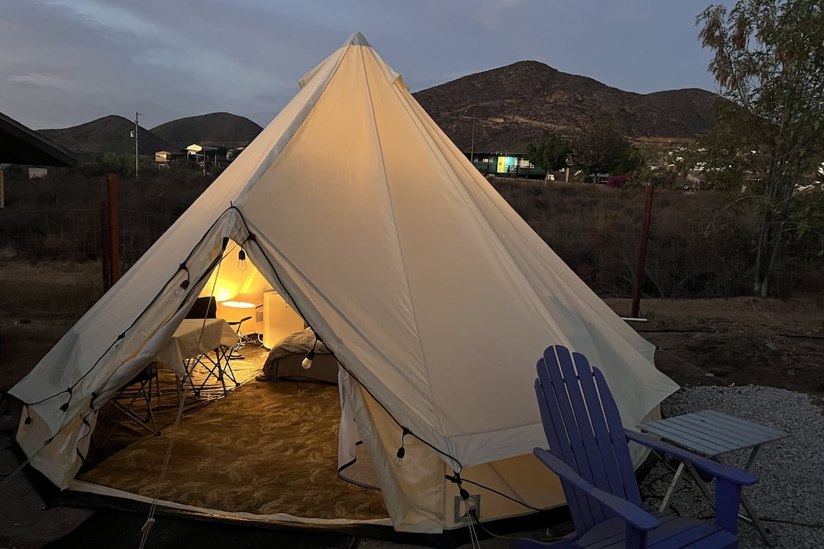 Uma tenda com mesa e cadeiras no interior, montada em um ambiente semelhante a um deserto, com montanhas ao fundo.