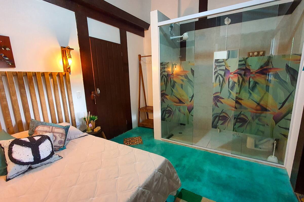 A room with a bed, a wooden slatted headboard, a glass shower enclosure with a floral design, and a green carpet.