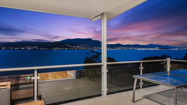 A balcony with a glass table and chairs, overlooking a coastal city at dusk with a view of the sea and distant mountains.