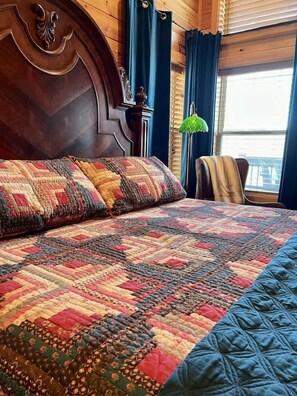 A bed with a quilt, a wooden headboard, a chair, and a window with curtains. - Jackalopia: Hot Tub, Firepit & Mountain Folklore!" (Gatlinburg)