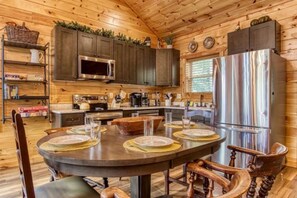 A wooden cabin interior with a dining table set for four, a kitchen with dark cabinets, a refrigerator, and a window with blinds.