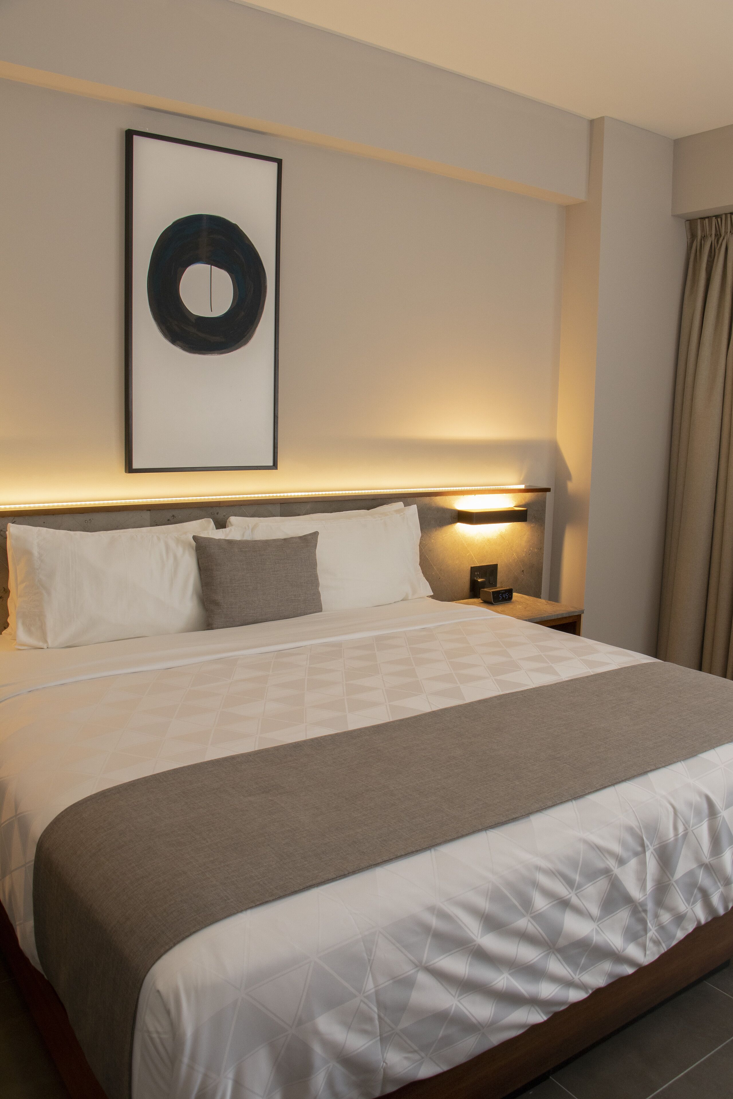 Een hotelkamer met een groot bed, een hoofdeinde met een lamp, een lijst met een afbeelding aan de muur en een nachtkastje met een lamp.