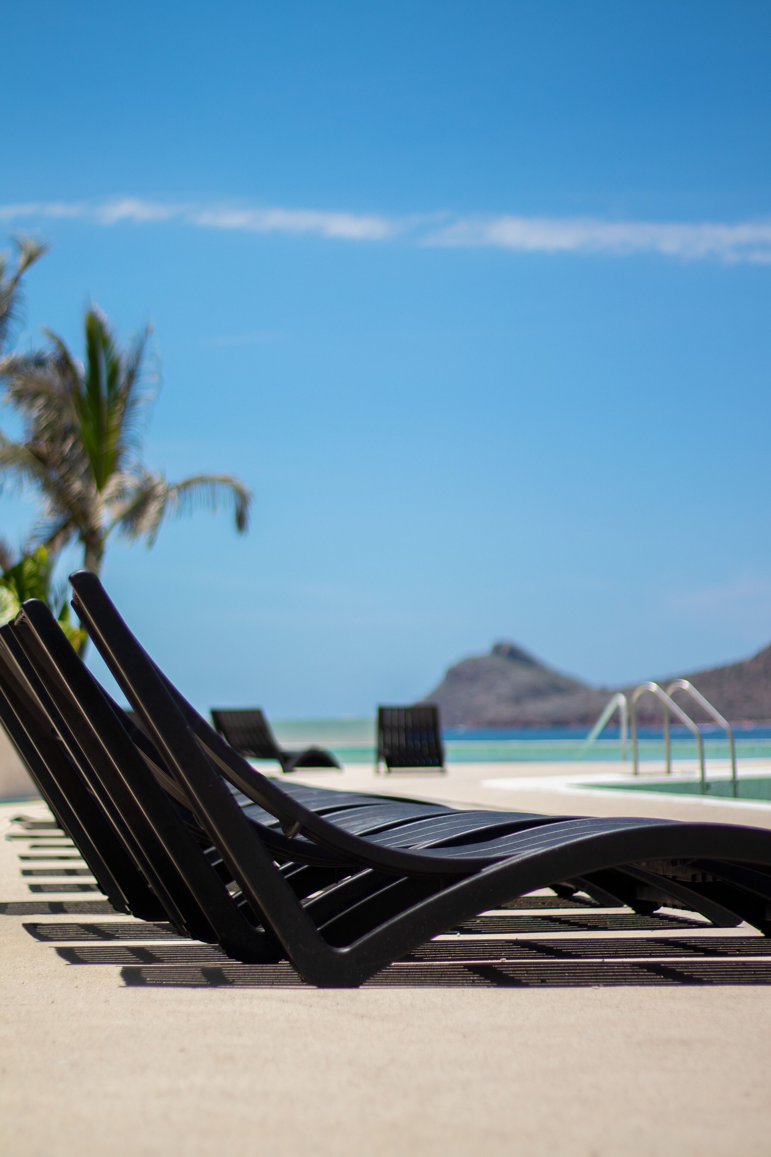 Un fauteuil de plage noir, sur une plage, avec un ciel bleu clair et des montagnes à l’horizon.