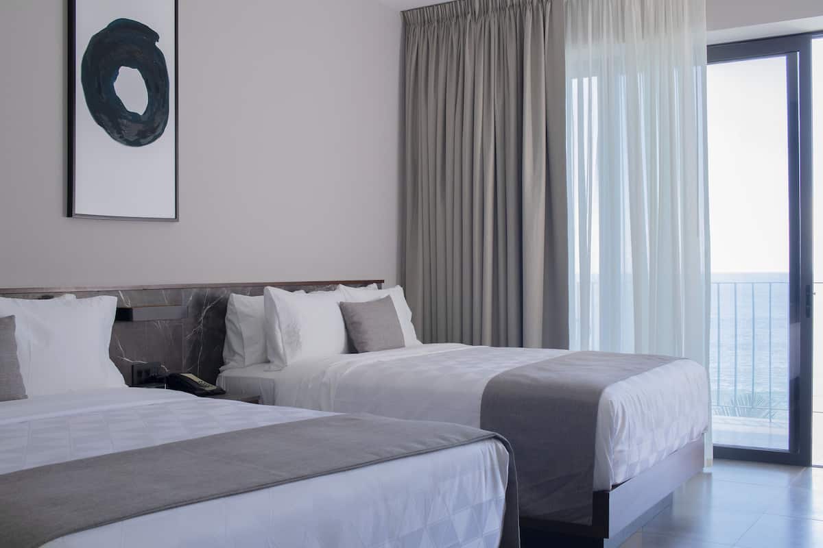 A hotel room with two beds, a large window with curtains, and a view of the sea.