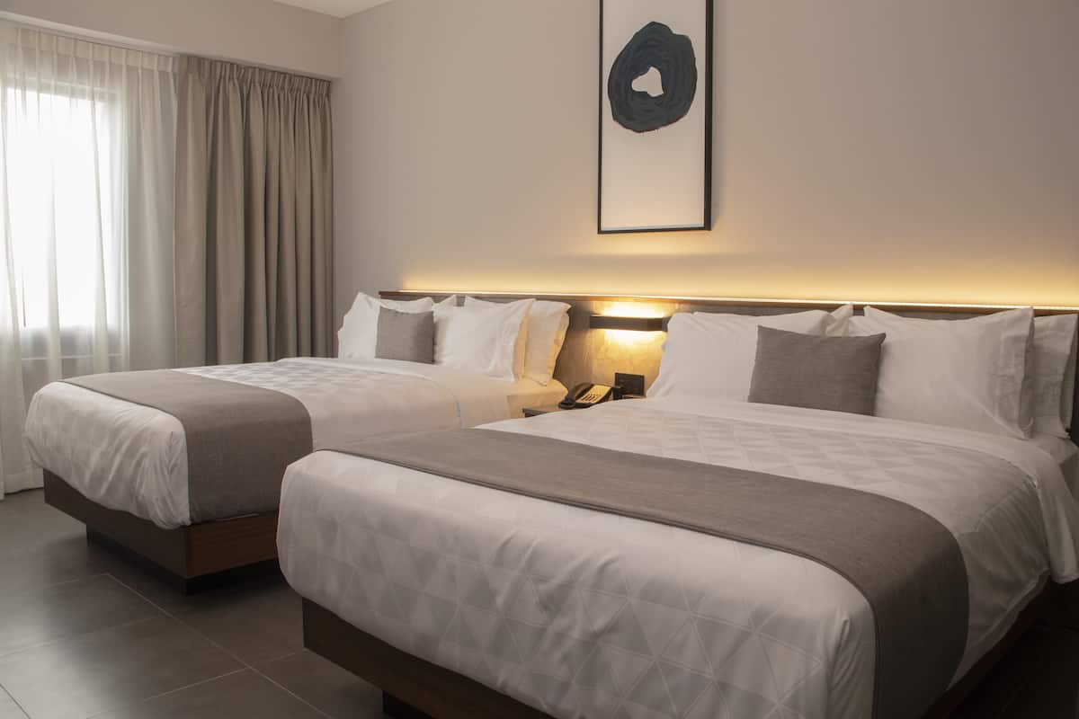 A hotel room with two beds, a bedside table with a lamp, and a framed artwork on the wall.