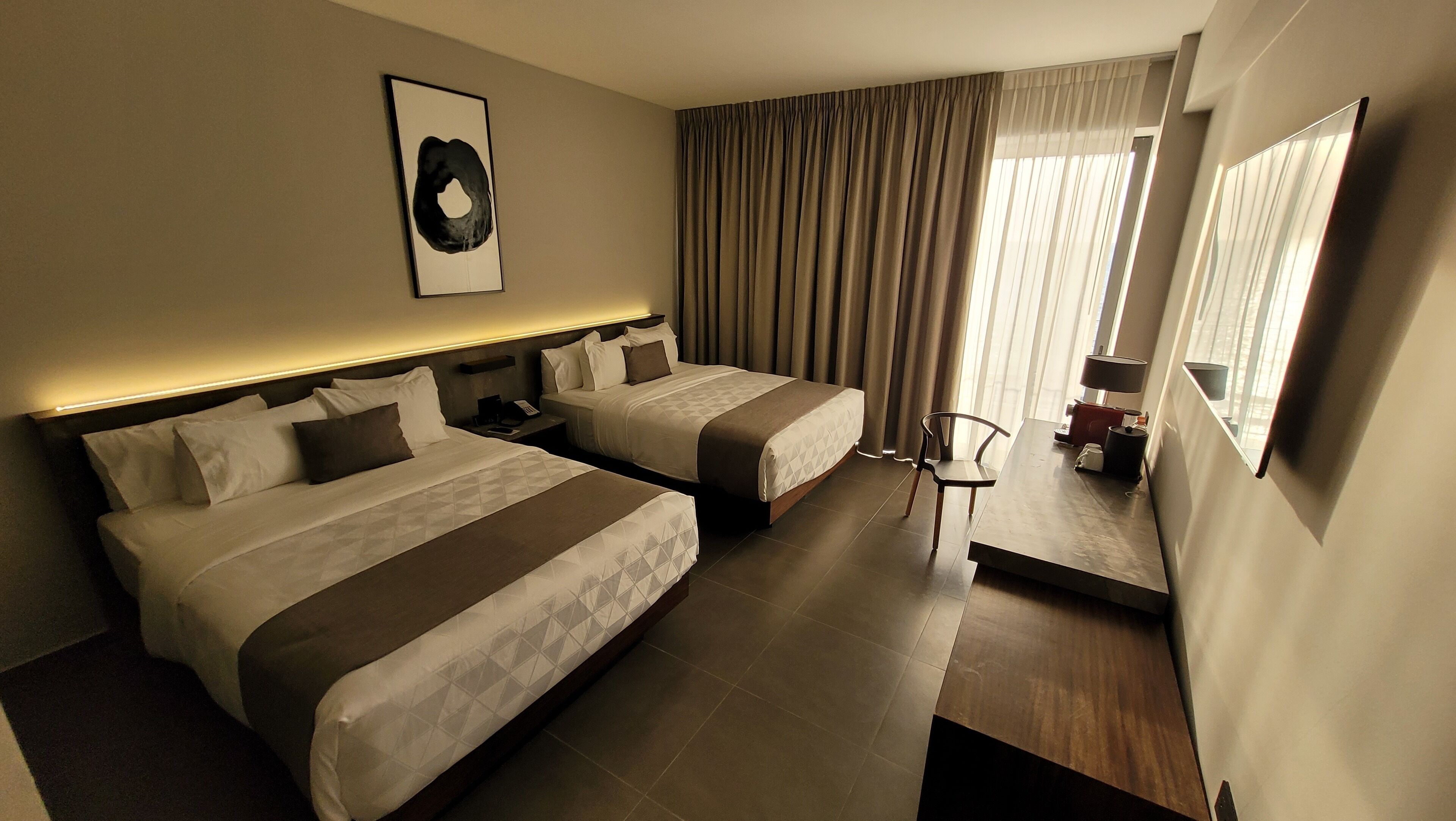 Une chambre d’hôtel avec deux lits, un bureau, une chaise, une lampe et une fenêtre avec des rideaux.
