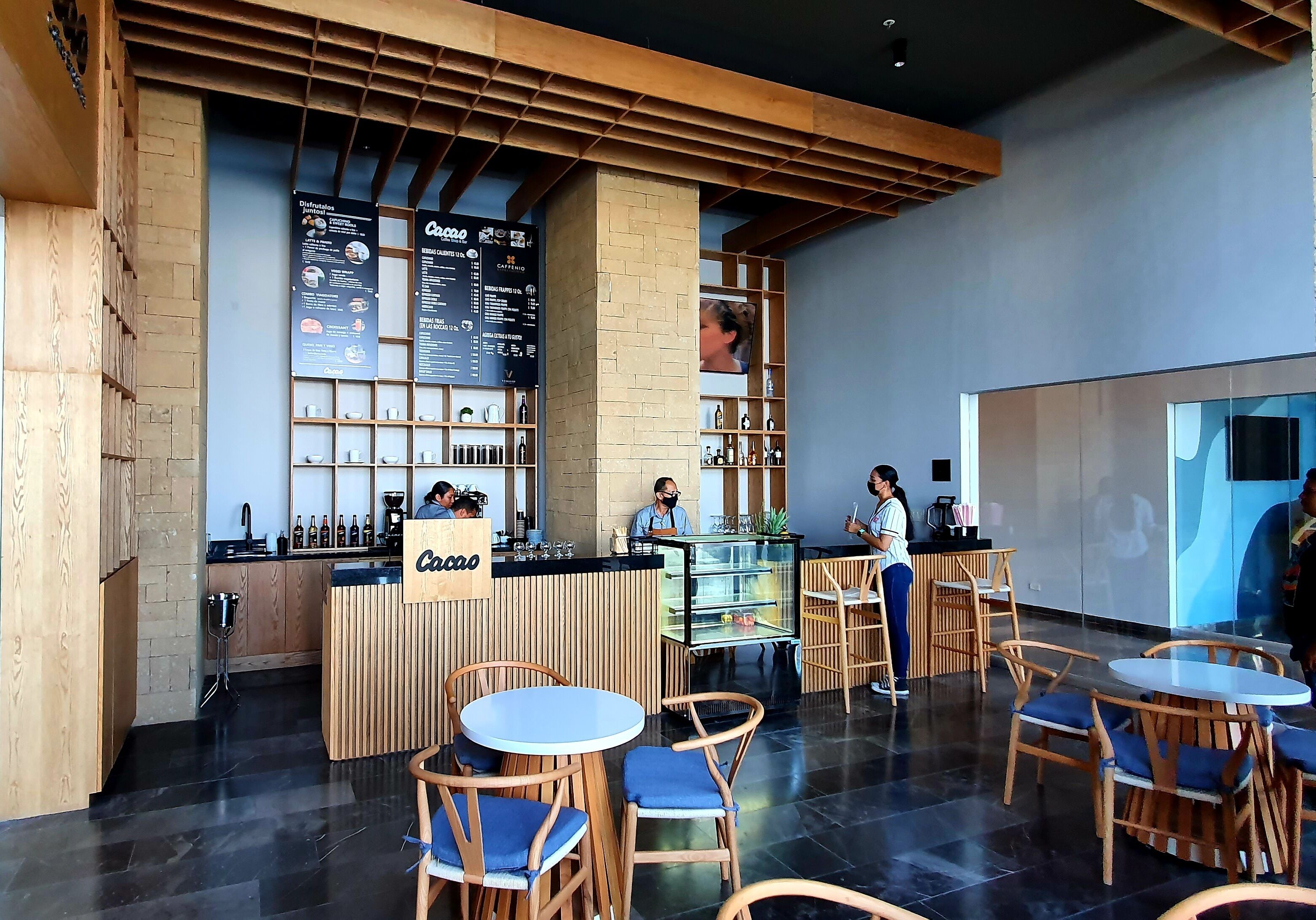 A modern cafe with wooden furniture, a counter with a menu, and a large screen displaying a person.
