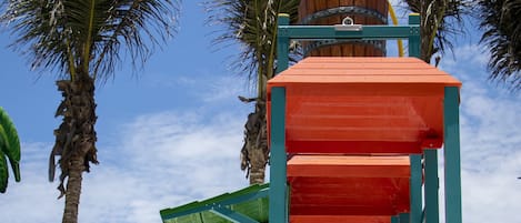 A playground with a slide and climbing structures, surrounded by palm trees and a clear sky.