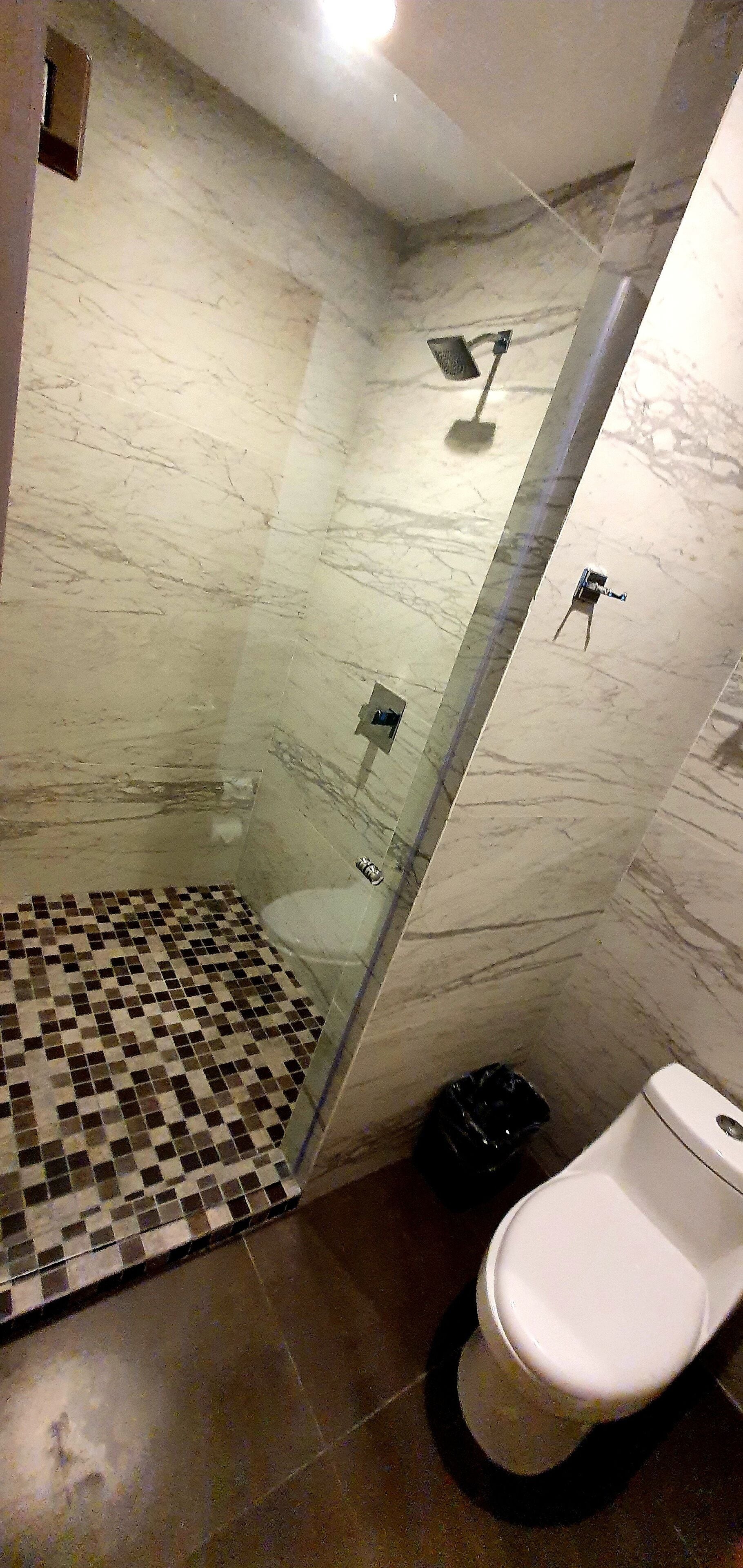 A bathroom with a glass-enclosed shower, a tiled floor, a toilet, and a trash bin.