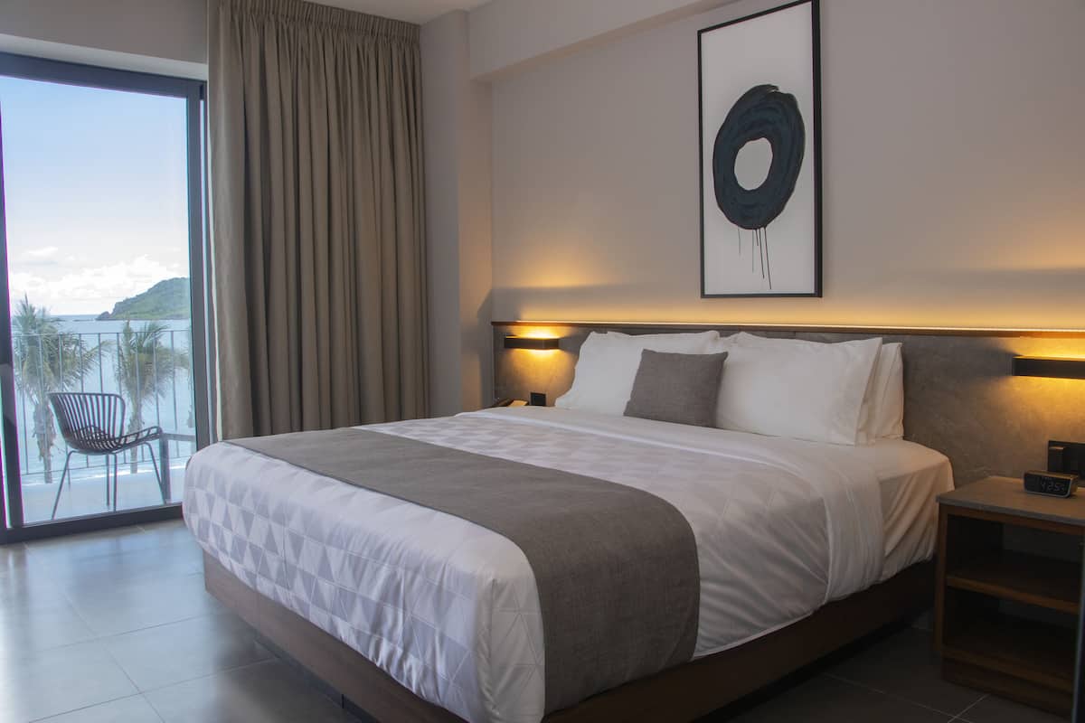 A hotel room with a large bed, a nightstand, a wall-mounted abstract painting, and a view of the sea through a sliding glass door.