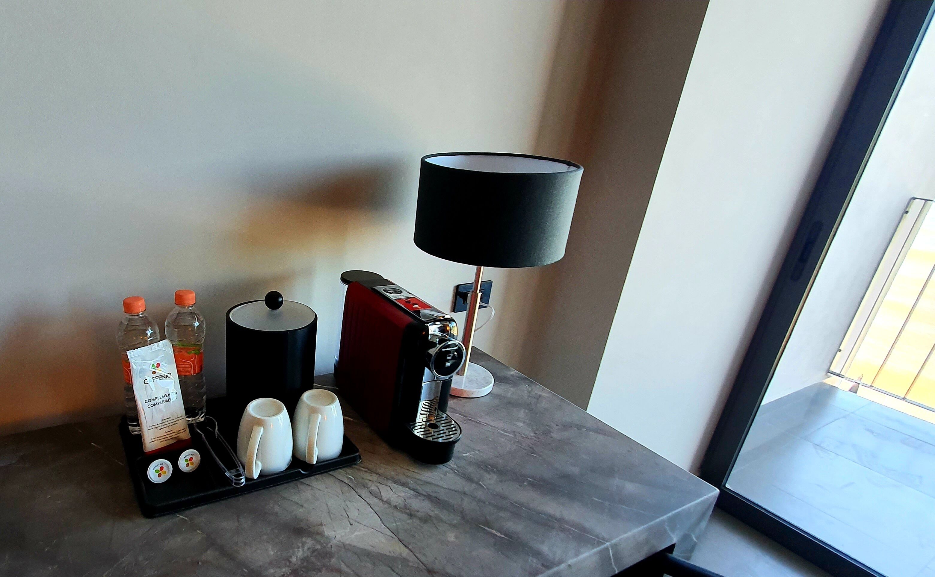 Una cafetera, dos tazas, una bandeja con dos botellas y un paquete, y una lámpara sobre una mesa cerca de una ventana.