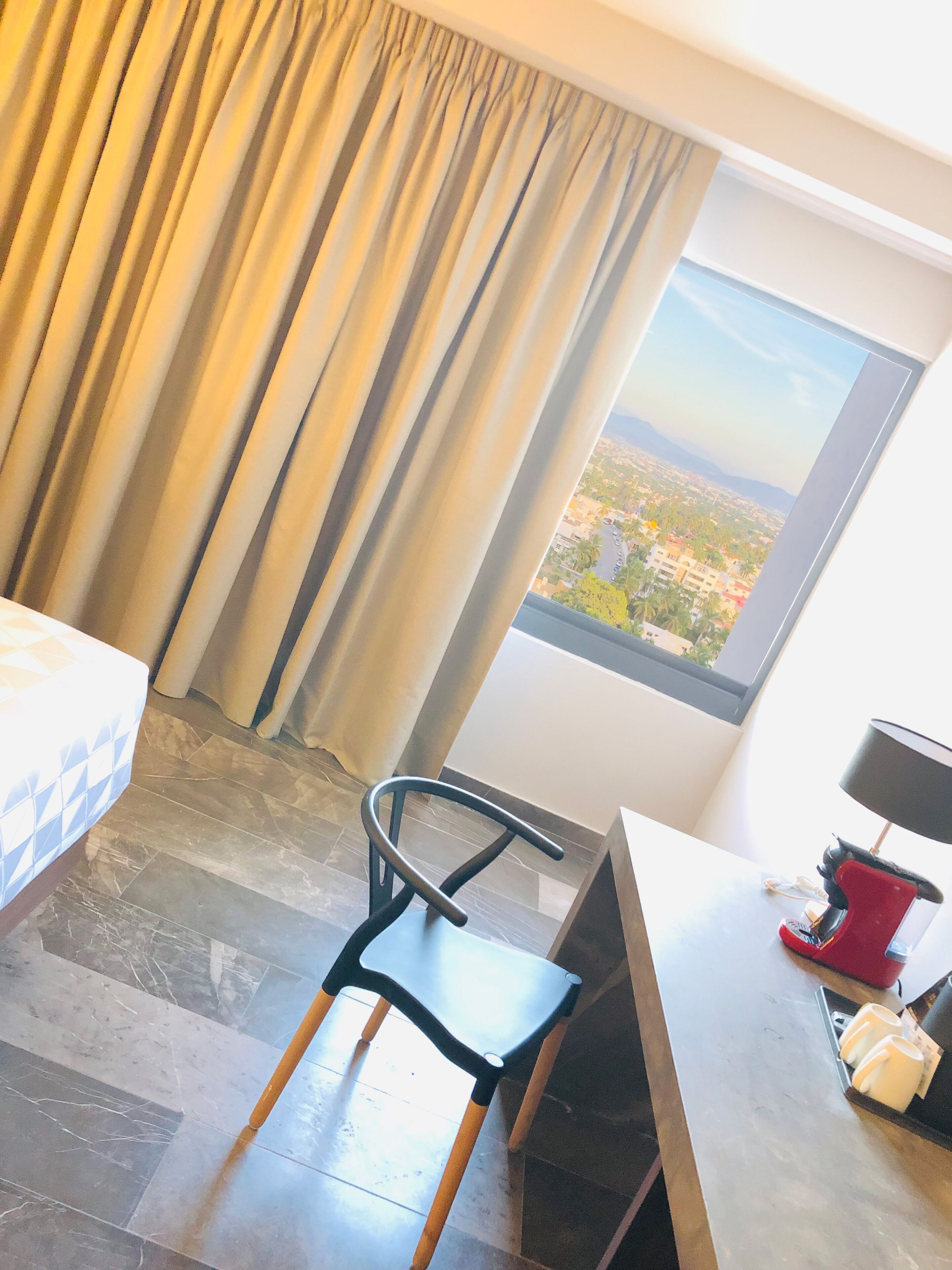 A hotel room with a bed, a chair, a desk, a lamp, and a window with a city view.
