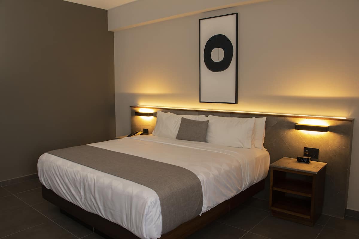 A modern hotel room with a large bed, bedside lamps, a framed artwork on the wall, and a small wooden nightstand.