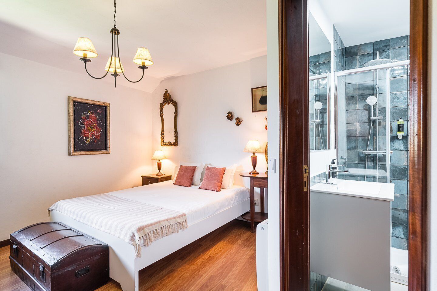 A bedroom with a bed, bedside lamps, a wooden chest, a mirror, and a window with a view of a bathroom.