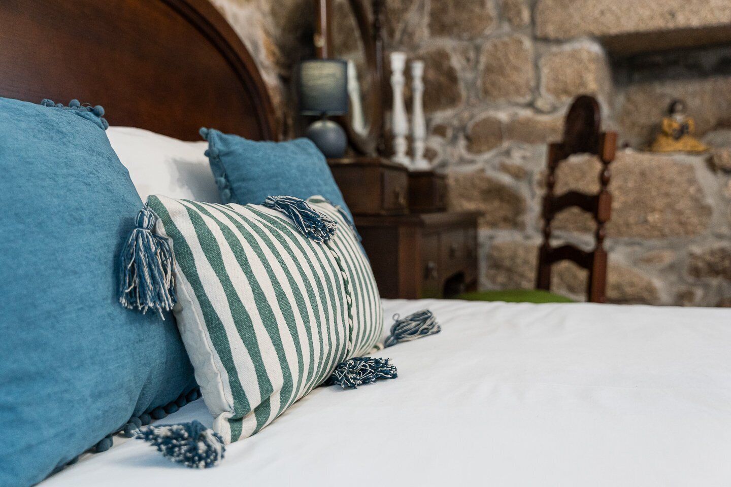 A bed with blue and striped pillows, a wooden headboard, and a stone wall background.