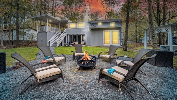 A modern outdoor seating area with a fire pit, surrounded by wicker chairs with cushions, set against a backdrop of a house and trees.