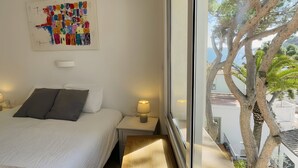 A bedroom with a bed, bedside tables, a wall art piece, and a large window with a view of trees and a building. - Stunning & Intimate views. Terrace + Rooftop. Wifi (Tamariu)