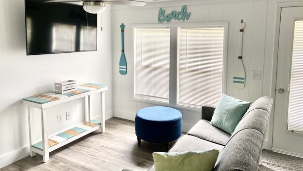 Een kamer met een witte wand, een plafondventilator, een televisie bevestigd boven een witte console tafel met een houten blad, en een blauwe poef.