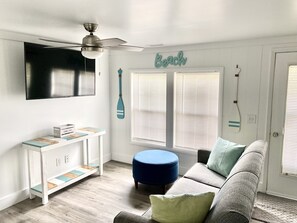 Una habitación con pared blanca, ventilador de techo, televisor montado sobre un mueble blanco con tapa de madera y un reposapiés azul.