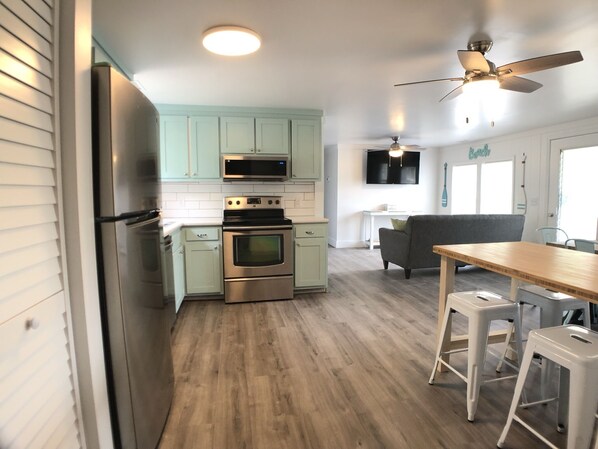Una cocina moderna con electrodomésticos de acero inoxidable, una zona de comedor con una mesa de madera y taburetes blancos, y un ventilador de techo.