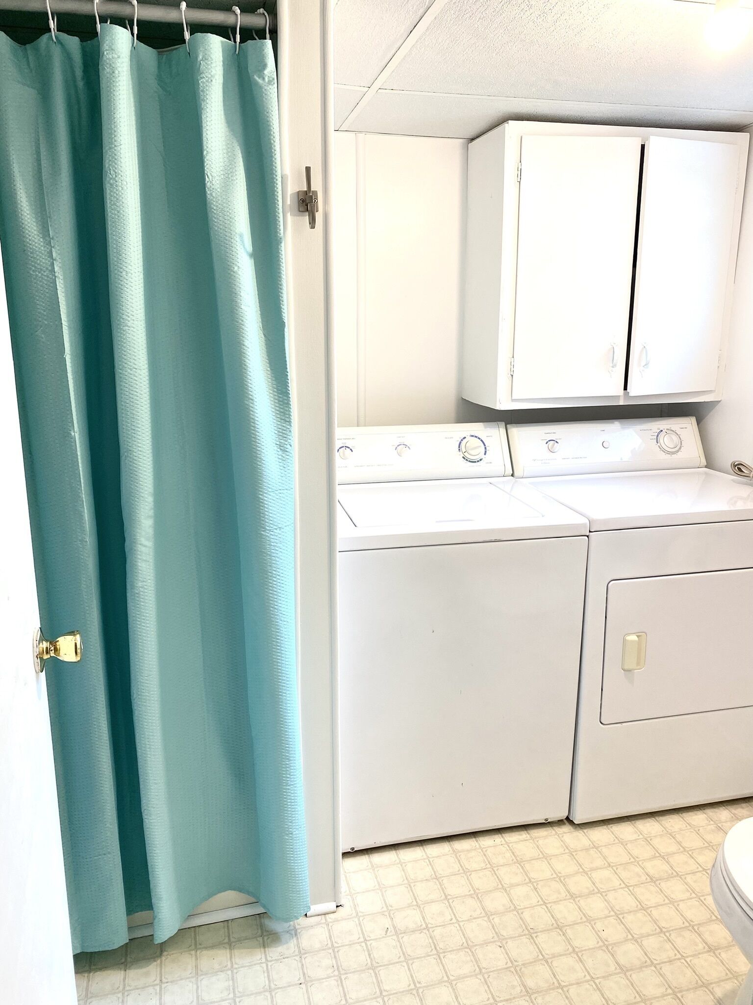 Un cuarto de lavandería con lavadora y secadora blancas, cortina de ducha color turquesa y piso cubierto de baldosas blancas.